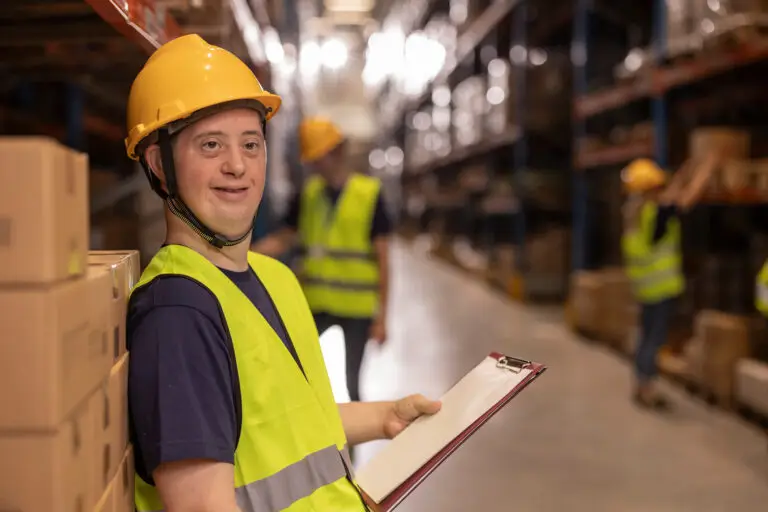 Young warehouse worker with down syndrome holding a clipboard and smiling confidently while colleagues engage in various tasks in the background, showcasing teamwork and inclusion in the workplace