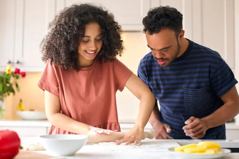 Couple At Home With Man With Down Syndrome And Woman Rolling Dough For Pizza In Kitchen Together