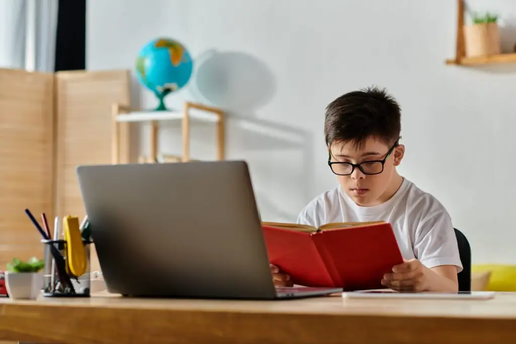 Young man in glasses reading a book at a desk with a laptop