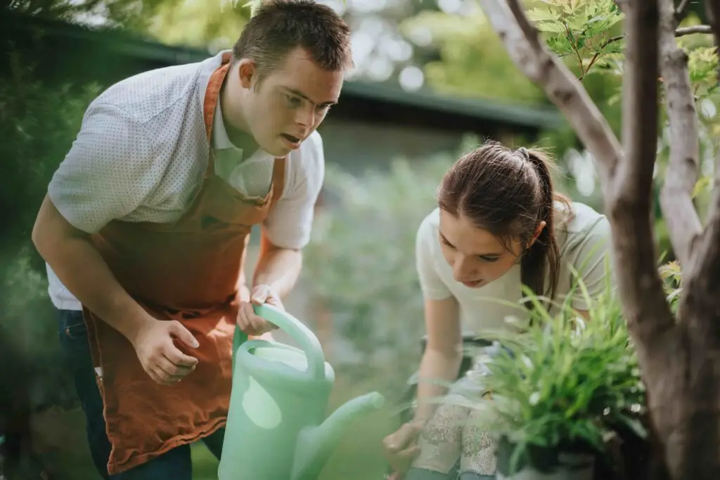 Man in an apron watering plants with a woman helping beside him